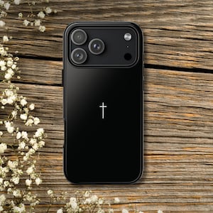 May include: Black phone case with a white cross design. The case has a glossy finish and features a triple-lens camera system. The background is a wooden surface with white flowers.