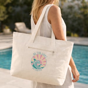 May include: A large off-white canvas tote bag featuring a zippered front pocket and a circular graphic print. The design displays a stylized ocean wave and sun rays in shades of teal, pink, and yellow, with the text BE THE LIGHT MATTHEW 5:14.