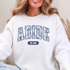 May include: White sweatshirt with the word "ABIDE" in blue floral print lettering. Below the word "ABIDE" is the phrase "IN HIM" in a blue oval. The sweatshirt is a crew neck style.