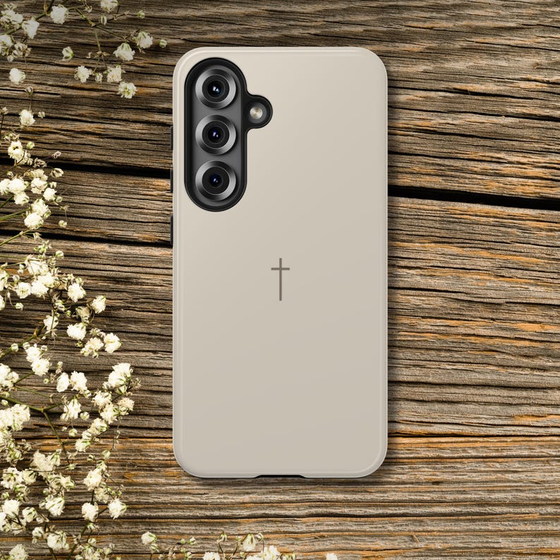 K&ouml;nnte beinhalten: Beige Handyh&uuml;lle mit einem Kreuzsymbol. Das Geh&auml;use hat eine Dreifach-Kameraanordnung oben links. Der Hintergrund ist eine Holzoberfl&auml;che mit wei&szlig;en Blumen.