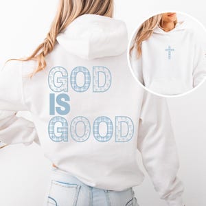 May include: White hoodie with the text "GOD IS GOOD" in a light blue plaid pattern on the back. A small light blue plaid cross is on the front. The hoodie has a drawstring hood and a front pocket.