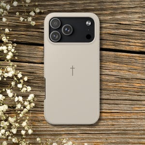 K&ouml;nnte beinhalten: Beige Handyh&uuml;lle mit einem Kreuzsymbol. Die H&uuml;lle hat ein sch&uuml;tzendes Design mit einem erh&ouml;hten Rand um die Kameralinsen. Der Hintergrund ist eine Holzoberfl&auml;che mit wei&szlig;en Blumen.