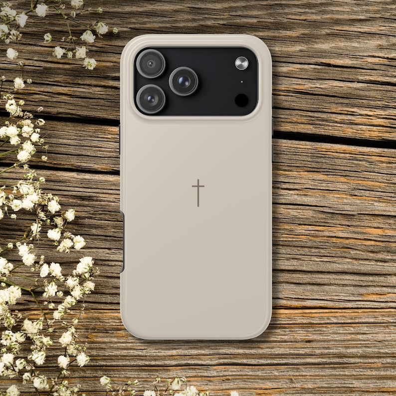 K&ouml;nnte beinhalten: Beige Handyh&uuml;lle mit einem Kreuzsymbol. Die H&uuml;lle hat ein sch&uuml;tzendes Design mit einem erh&ouml;hten Rand um die Kameralinsen. Der Hintergrund ist eine Holzoberfl&auml;che mit wei&szlig;en Blumen.