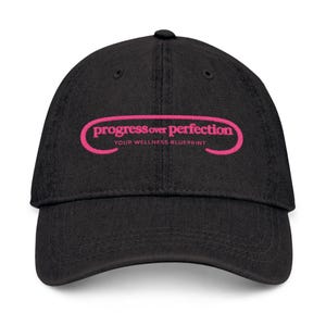 Peut inclure: Casquette de baseball noire avec une visière incurvée. Le chapeau affiche le texte rose "progress over perfection" dans une police stylisée, avec les mots "YOUR WELLNESS BLUEPRINT" en dessous.