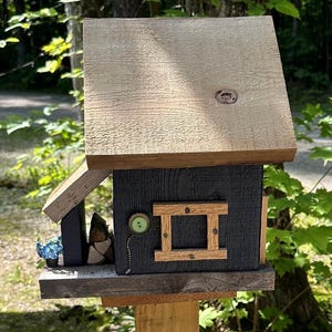 May include: A handcrafted wooden birdhouse with a light brown roof and a dark gray body. The birdhouse features a decorative window and a green button detail. It is mounted on a wooden post and set against a natural outdoor backdrop.
