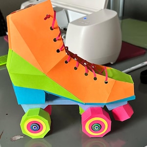Puede incluir: Un patín de papel colorido en tonos naranja, verde, azul y rosa. El patín presenta cordones rosas y ruedas con círculos concéntricos. El diseño es geométrico y angular, con una estética lúdica y vibrante.