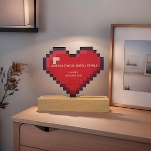 Op de afbeelding: Een gepixelde rode lamp in de vorm van een hart met de tekst "LOVE.EXE STATUS: MOSTLY STABLE" en "PLAYER 2 UNLOCKED 2018" op een houten voet.