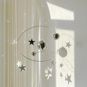 Könnte beinhalten: Ein silbernes Mobile mit einer Discokugel, Sternen und einem weißen Hintergrund. Das Mobile hängt von der Decke.