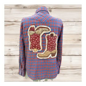 May include: A long-sleeved plaid shirt in red and blue, featuring appliqued cowboy boots with star patterns. The boots are in shades of brown and red. The shirt is displayed on a mannequin.