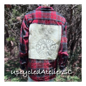 May include: Red and black plaid flannel shirt with a beige patch on the back. The patch features a circular design with the words "FULTON SEAMLESS EXTRA HEAVY" and an eagle graphic. The shirt is button-down and has long sleeves.