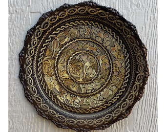 May include: A decorative, round, dark brown plate with intricate floral and geometric patterns in gold and bronze tones. The plate has a scalloped edge and is displayed against a white wooden background. The plate's design is centered with concentric circles.