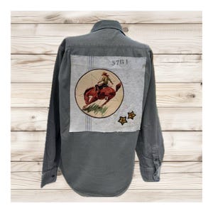 May include: Gray long-sleeve shirt with a patch featuring a rodeo rider on a bucking bronco. The patch is circular with a brown horse and rider, surrounded by a light gray background. Two star patches are below the circular patch, and the number "5761" is above it.