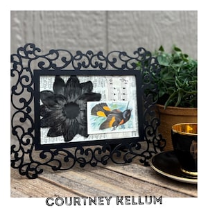 May include: A black decorative picture frame with an ornate, swirling design. The frame holds a black flower, a small bird illustration, and sheet music. A black teacup with gold trim sits nearby. The text "COURTNEY KELLUM" is at the bottom.