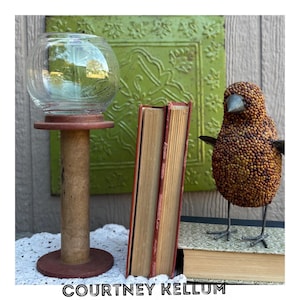 May include: A clear glass bowl sits atop a wooden stand with a red base. Two vintage books with red spines stand next to the stand. A decorative bird made of small beads is also present. The text "COURTNEY KELLUM" is at the bottom.