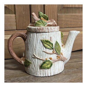 May include: A ceramic teapot shaped like a white tree stump with a textured bark finish. The handle and spout resemble brown branches. The lid features a sculpted branch with green leaves and small orange berries, matching the branch design on the body.