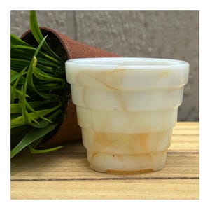 May include: A small, tiered planter featuring a translucent, milky white and amber marbled glass finish. The vessel has a stepped, circular profile with a wide rim and a narrow base, showcasing a smooth, polished surface with swirling patterns.