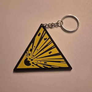 Può includere: Un portachiavi giallo a forma di triangolo con una grafica di esplosione nera. Il portachiavi ha un anello di metallo argentato.