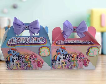 May include: Two decorated gift boxes, one blue and one pink, featuring cartoon ponies. Each box has a purple bow with a gem, the number 3, and the name "CAMILLE" on top. The boxes are on a wooden surface.