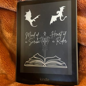 May include: A black and white Kindle e-reader with the text "Mind of a Scribe, Heart of a Rider" on the screen. The screen also features a stylized image of a book, a dagger, and two dragons.