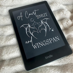 Op de afbeelding: Een zwart-witte Kindle e-reader met de tekst "A Court SMUT and WINGSPAN" op het scherm. Het Kindle-logo is zichtbaar onderaan het scherm.