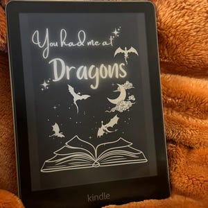 Puede incluir: Un lector electrónico Kindle en blanco y negro con el texto "You had me at Dragons" en la pantalla. La pantalla también presenta una imagen estilizada de un libro, una luna creciente y varios dragones.