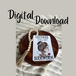 Op de afbeelding: Een witte kaart met de tekst "Tattooed, Introverted, Bookworm" en een kleurrijke illustratie van een vrouw met een boekenkast als haar. De kaart ligt op een houten dienblad met een houten kralensnoer.