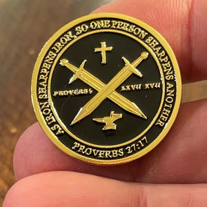 May include: Gold-colored metal coin with a black background. The coin features two crossed swords, a small anvil, and a cross. The text "As iron sharpens iron so one person sharpens another" is written around the edge of the coin. The text "Proverbs 27:17" is written at the bottom of the coin.
