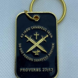 May include: Gold-colored keychain with a black rectangular tag. The tag features a gold-colored cross and two swords crossed over a gold-colored anvil. The text on the tag reads "As iron sharpens iron, so one person sharpens another" and "Proverbs 27:17".