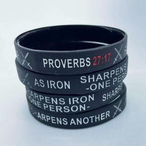 May include: A stack of black silicone bracelets with white text that reads "Proverbs 27:17 As Iron Sharpens -One Person- Sharpens Another" with a crossed sword graphic on each bracelet.