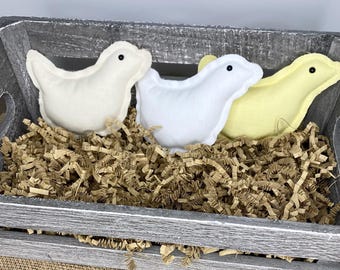 May include: Three felt bird-shaped toys in a wooden crate filled with brown shredded paper. The birds are white, yellow, and cream colored.