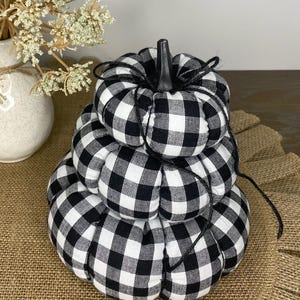 May include: A decorative, stacked pumpkin in black and white buffalo check fabric. The pumpkin has a black stem and is tied with black twine. The pumpkin is a fall-themed decoration.