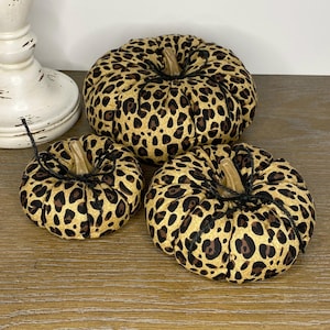 May include: Three decorative pumpkins with a leopard print pattern. The pumpkins have a tan background with brown spots and a brown stem. The pumpkins are different sizes and are arranged on a wooden surface.