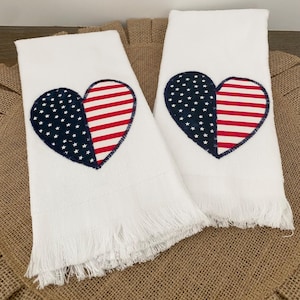 May include: Two white hand towels with a patriotic heart design. Each heart is split, with one half featuring a blue field of white stars and the other half displaying red and white stripes. The towels have a fringed edge.