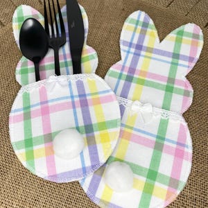 May include: Two pastel plaid bunny-shaped fabric place card holders with white cotton tails. Each holder has a white lace trim and a white bow. The holder on the left has a fork, knife, and spoon inside.