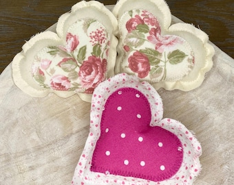 May include: Three fabric heart-shaped pillows. Two are white with a floral pattern and one is pink with white polka dots.