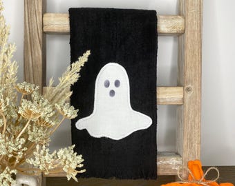 以下が含まれることがあります： 白いお化けのアップリケが付いた黒いハロウィーンをテーマにしたハンドタオル。お化けには3つの灰色の目の穴があります。タオルは、装飾的なオレンジ色のカボチャと一緒に木製の梯子に飾られています。