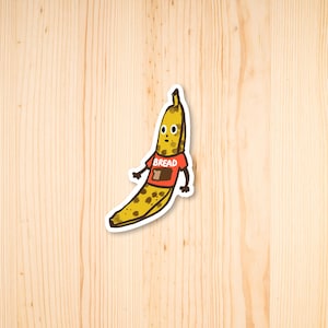 Könnte beinhalten: Eine Cartoon-Banane mit einem roten Shirt, auf dem "Brot" steht und ein Stück Brot darauf. Die Banane ist gelb mit braunen Flecken.