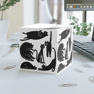 Puede incluir: Bloc de notas blanco en forma de cubo con siluetas negras de gatos en varias poses. El bloc de notas tiene un diseño en blanco y negro.