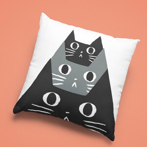 May include: A white pillow with a black and gray cat design. The cats are stacked on top of each other, with the largest cat at the bottom and the smallest cat at the top. The cats have simple, cartoon-like faces.