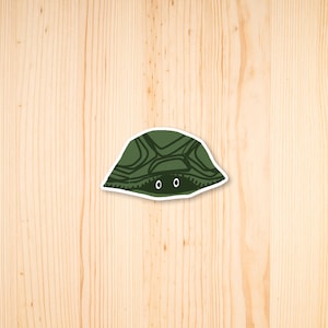 May include: A sticker featuring a green turtle shell with a patterned design. The turtle's eyes peek out from beneath the shell. The sticker is on a light wood grain background.