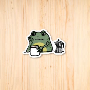 May include: A green frog cartoon illustration with a cup of coffee and a coffee pot. The frog is sitting at a table and holding the cup of coffee in its hand.