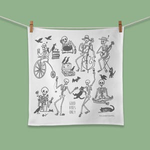 May include: A white tea towel with a black and white illustration of skeletons playing musical instruments, riding a bicycle, and holding drinks. The text "Good Vibes Only" is printed on the towel.