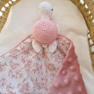 Peut inclure: Une couverture de sécurité pour bébé faite à la main avec une tête et une carapace de tortue crochetées en blanc et rose. La couverture a un imprimé floral d'un côté et un tissu rose doux et texturé de l'autre.