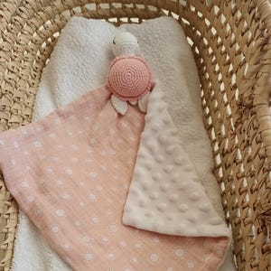 Peut inclure: Une couverture de confort pour bébé avec une tête de tortue crochetée et un tissu à motifs roses et blancs. La couverture a un envers blanc doux et texturé et est placée dans un panier en osier.