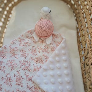 Puede incluir: Una manta de seguridad para bebés con una cabeza de tortuga de ganchillo y un caparazón rosa. La manta presenta un estampado floral en tonos rosas y blancos por un lado y un tejido suave y blanco con textura por el otro. La manta está dentro de una cesta tejida.