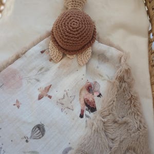 Peut inclure: Une couverture de sécurité pour bébé faite à la main, avec une tête et une carapace de tortue crochetées en marron et beige. La couverture a un tissu à motifs avec des illustrations d'oiseaux, d'étoiles et de montgolfières, et une bordure en fausse fourrure.