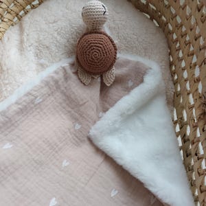 Peut inclure: Une couverture de sécurité en mousseline beige avec une petite tortue crochetée attachée à un coin. La couverture possède un envers en fausse fourrure blanche et un devant beige clair orné d'un motif répétitif de petits cœurs blancs imprimés.