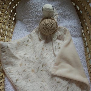 Peut inclure: Une couverture de sécurité pour bébé avec une tête et une carapace de tortue crochetées en beige et crème. La couverture présente un motif floral aux tons neutres doux et un coin en peluche de couleur pêche. La couverture est posée sur une serviette blanche.