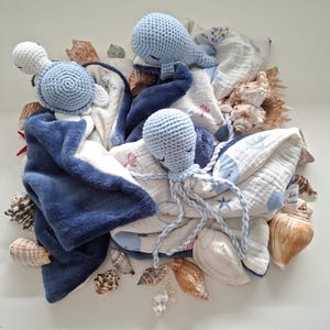 Peut inclure: Deux doudous au crochet représentant une baleine bleu clair et une pieuvre bleu clair avec de longs tentacules tressés. Les couvertures sont en tissu blanc avec un motif marin bleu et un envers en peluche bleu foncé, entourées de coquillages.