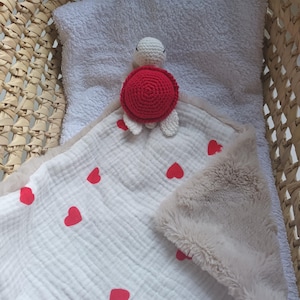 Puede incluir: Una manta de bebé blanca con patrones de corazones rojos y un borde de felpa beige. Una tortuga de ganchillo hecha a mano con caparazón rojo y cabeza y patas blancas está sobre la manta. La manta está dentro de una cesta de mimbre.